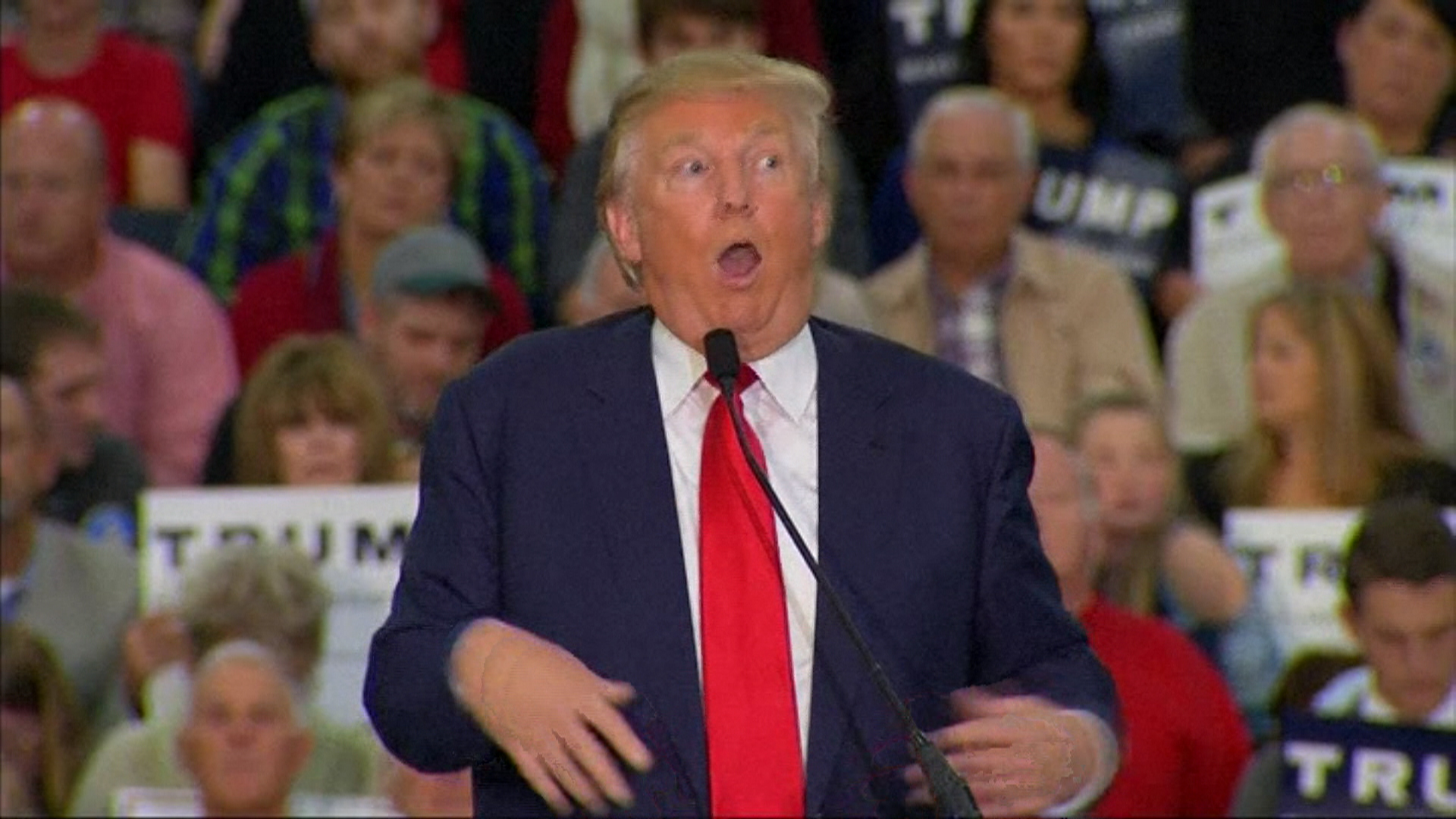 Donald Trump standing at a podium in front of a crowd, making exaggerated body movements and facial expressions with his mouth open wide.