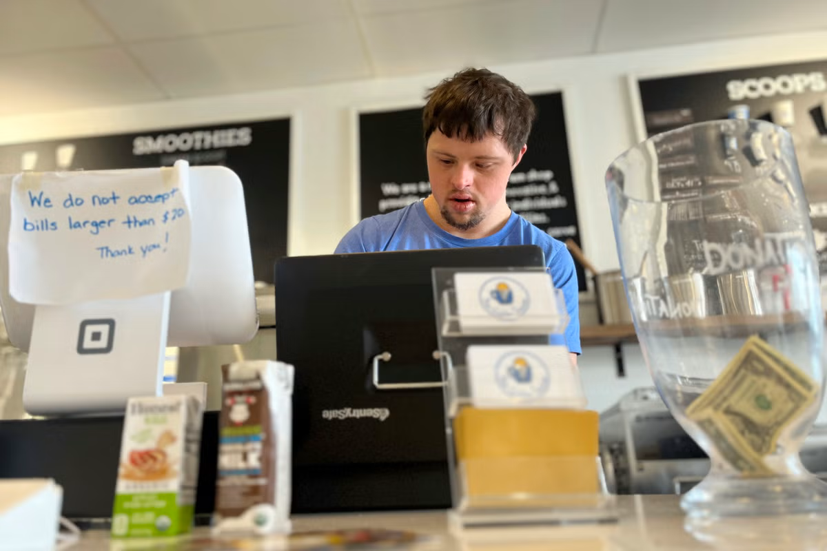 A person working at a counter in a café named "Scoops & Smoothies," standing next to a cash register and various beverages,