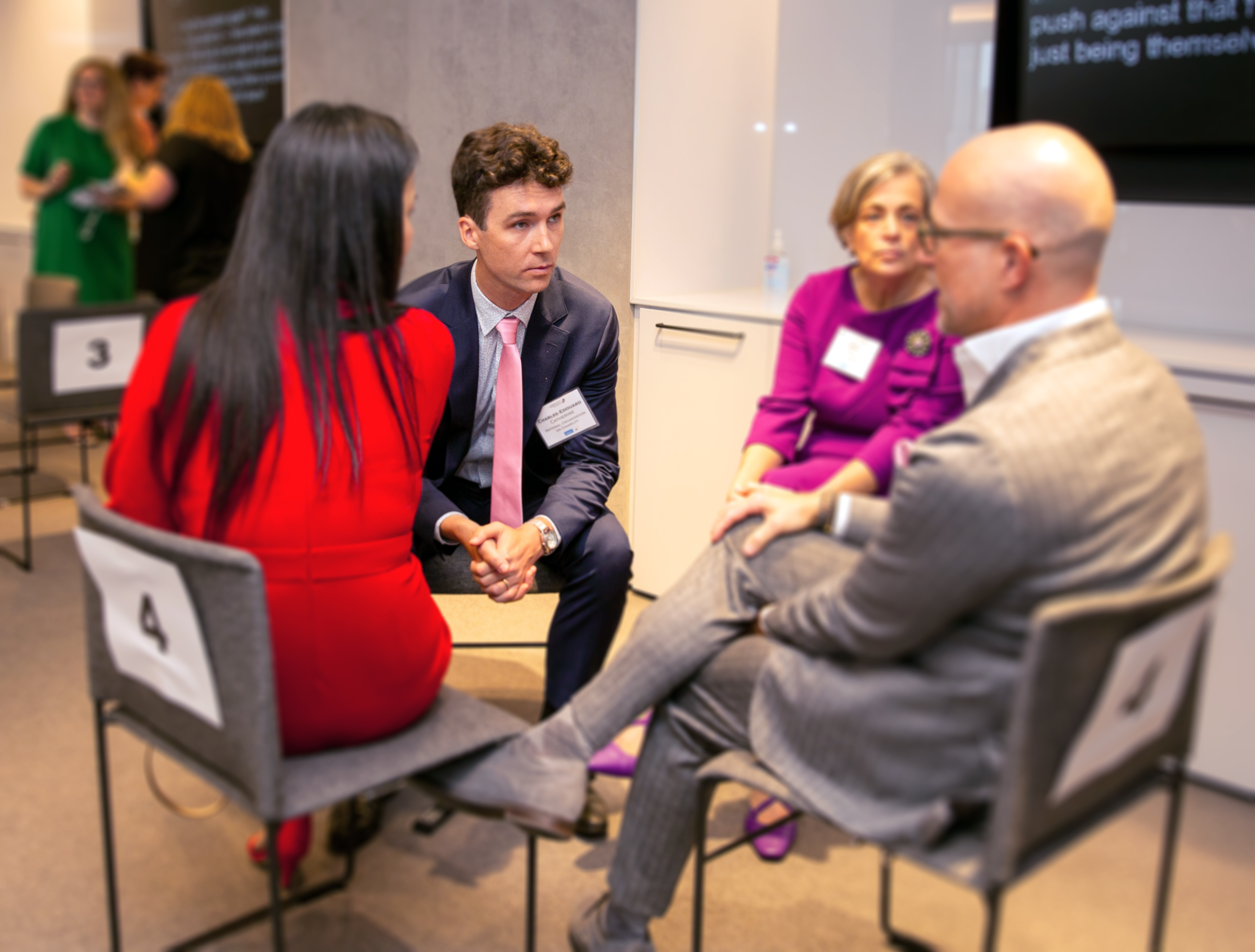 A group of four individuals seated in a circle engaged in discussion. From left to right: Margaret Ling, Charles Edaurd Catherine-Caldaro, Beth Sirull, and Shawn VanDerziel. The background shows blurred activity, including people standing and a screen displaying text. The setting appears to be a professional event or workshop.