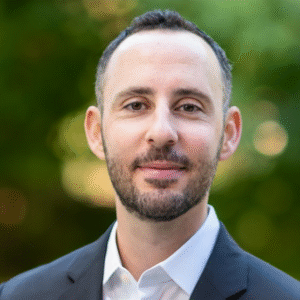 Dan Robin Headshot for Dan Robin, a white man with dark hair and a beard looking directly at the camera and smiling. He wears a white shirt and navy blazer. The background is blurred, but it appears to be trees.