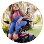Photo of woman with long brown hair sitting in a power wheelchair outside, smiling and looking off to the distance