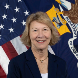 A blonde woman wearing a navy blazer smiling with an American flag in the background