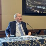 Congressman Jim Langevin sits in a power wheelchair speaks at a table with a microphone in front of him inside a conference room. He is wearing a dark suit and blue tie and is positioned in front of a screen displaying the "United States House of Representatives" logo with a background image of the Capitol at night. Papers are spread on the table, which is covered with a blue and gold patterned tablecloth.