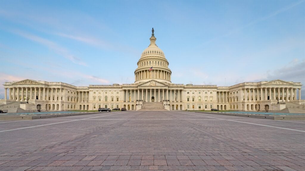 Wide-angle view of the entire United States Capitol Building in front of a bright, light blue sky with faint pink clouds.