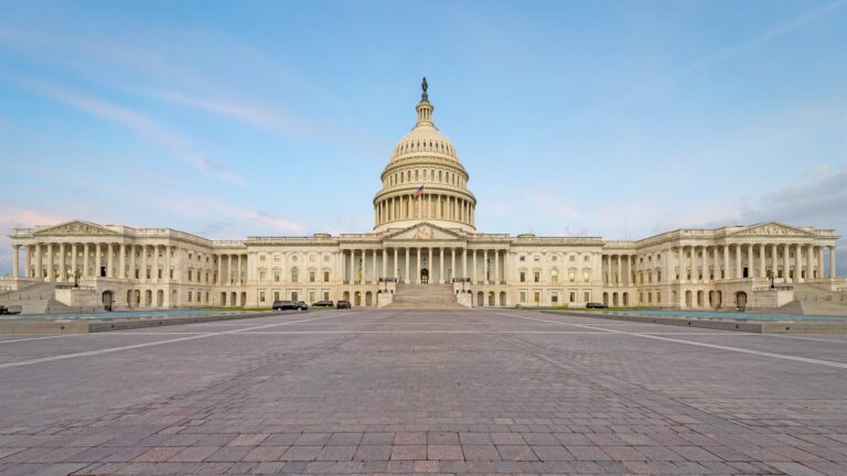 Wide-angle view of the entire United States Capitol Building in front of a bright, light blue sky with faint pink clouds.