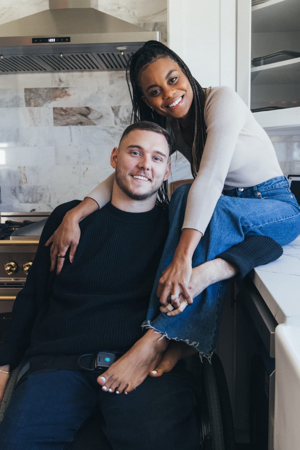 A woman sits on a ledge with her arms and legs wrapped around the man beside her, who is sitting in a wheelchair. Both are looking at the camera smiling.