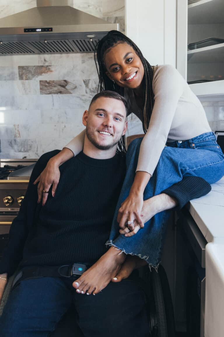 A woman sits on a ledge with her arms and legs wrapped around the man beside her, who is sitting in a wheelchair. Both are looking at the camera smiling.