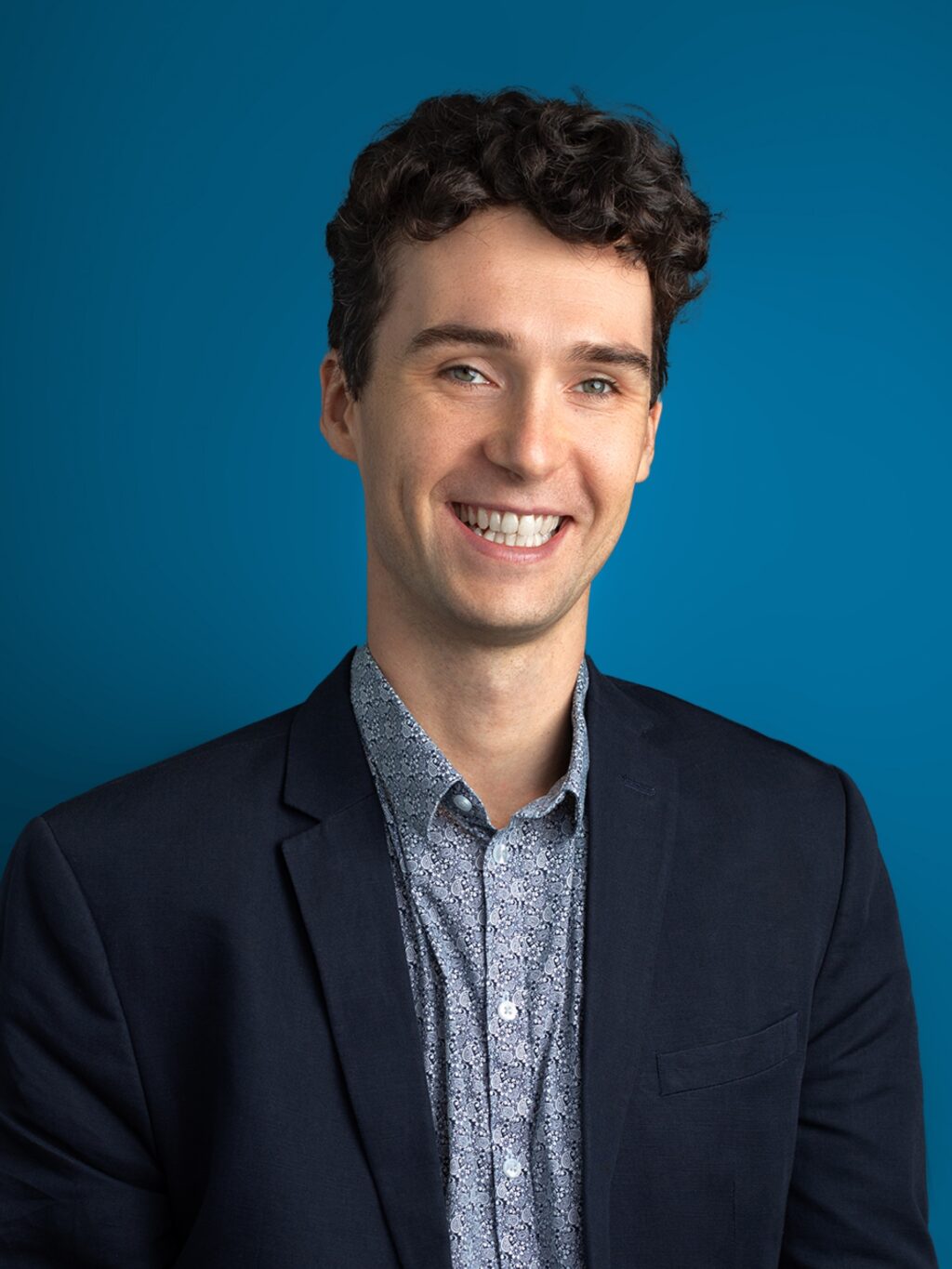 Smiling man with curly hair wearing a navy blazer and patterned shirt, set against a solid blue background. The tone is professional and friendly.