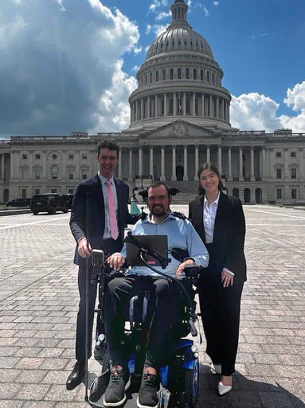 Three people pose in front of the U.S. Capitol building on a sunny day with dramatic clouds in the sky. A man in a power wheelchair is centered between two standing individuals in business attire. The wheelchair user is wearing a light blue shirt and black pants, and has assistive technology mounted on the chair. The group appears cheerful, and the scene suggests a visit or advocacy event on Capitol Hill. (From left to right: Charles-Edouard Catherine Vice President, Corporate & Government Relations, advocate Robby Barbieri, and Paulina Romero, Senior Manager, Partnerships, NOD)