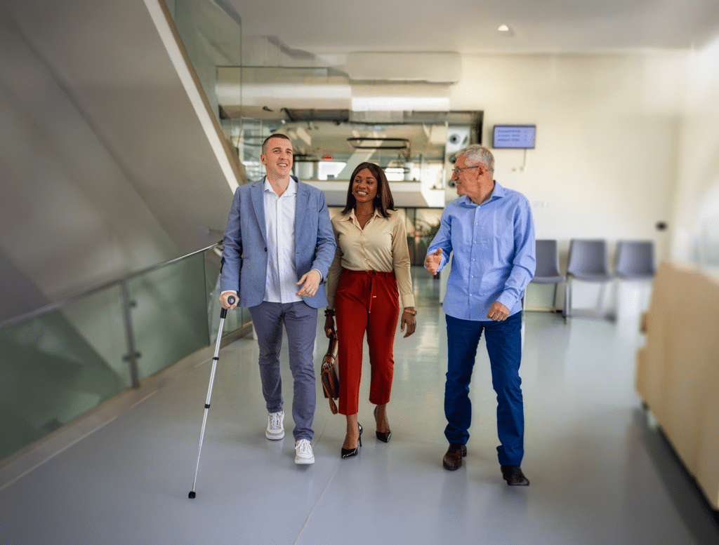 Three professionals walk side by side through a bright, modern office building. A man on the left uses a white cane, while the woman in the center and the older man on the right walk alongside him, smiling and talking as they move down the hallway.