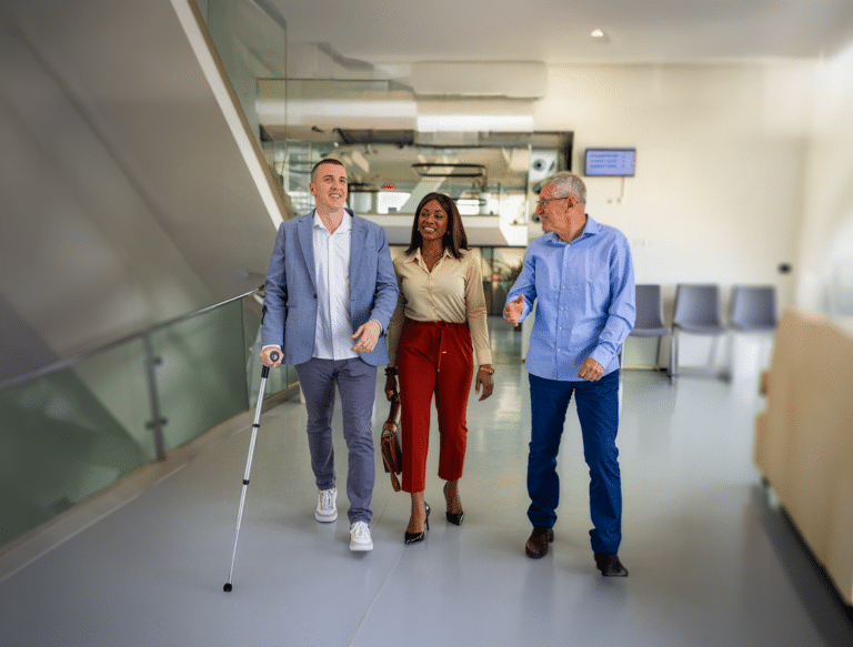 Three professionals walk side by side through a bright, modern office building. A man on the left uses a white cane, while the woman in the center and the older man on the right walk alongside him, smiling and talking as they move down the hallway.