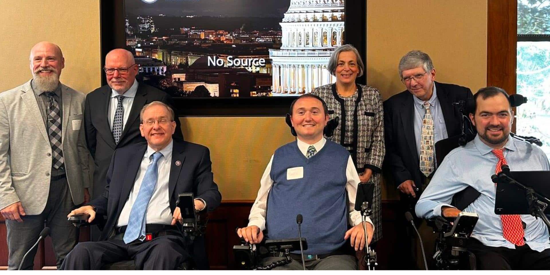 A group of seven people pose for a photo in a conference room at the U.S. House of Representatives. Three individuals are in power wheelchairs, while the others stand behind them. Everyone is smiling and dressed in business or professional attire. Behind the group is a large screen displaying an image of the U.S. Capitol and the House of Representatives logo. The setting suggests a formal meeting or advocacy event. (From left to right: Doug Crandell, Jim Calbi, Congressman Langevin, Josh Basile, Beth, Dr. Tony Antosh, Robby Barbieri)