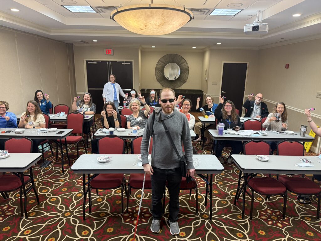 Matthew standing in front of a seated group of people, whose arms are outstretched holding Legos