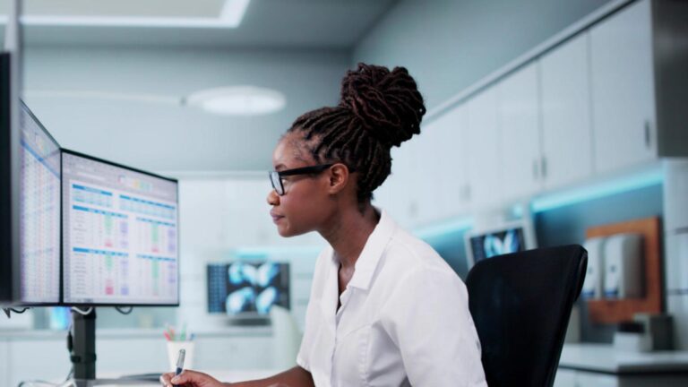 A woman in a white lab coat and glasses focuses intently on dual monitors displaying data in a modern lab. The setting conveys professionalism and focus.