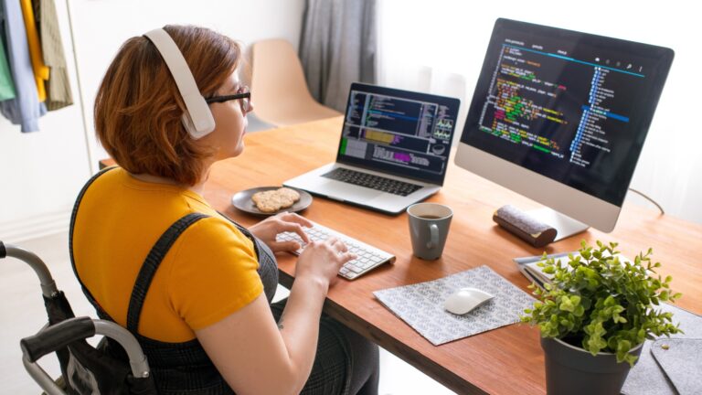 A woman with glasses in a wheelchair using her hands at a desk with a laptop. She's seated in an office, conveying a professional and focused atmosphere.