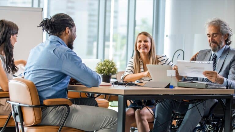 Four business professionals having a meeting around conference table in modern office with large windows. One team member uses a wheelchair, demonstrating workplace inclusion."Detailed version: Medium version: "Four business professionals having a meeting around conference table in modern office with large windows. One team member uses a wheelchair, demonstrating workplace inclusion.