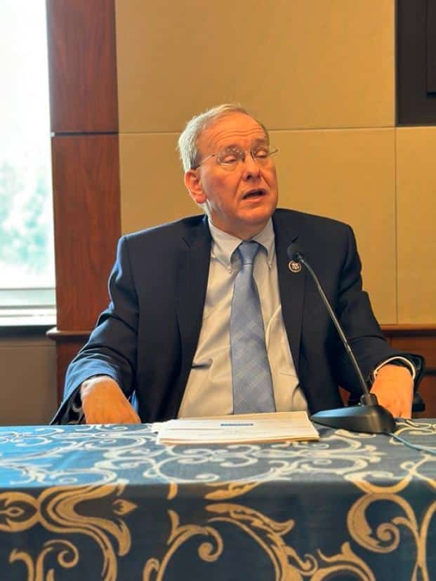 Congressman Jim Langevin sits in a power wheelchair and speaks at a table with a microphone in front of him inside a conference room. He is wearing a dark suit and blue tie and is positioned in front of a screen displaying the "United States House of Representatives" logo with a background image of the Capitol at night. Papers are spread on the table, which is covered with a blue and gold patterned tablecloth.