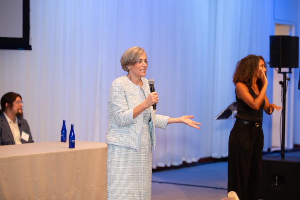 A woman holding a microphone addressing a crowd. Beside her stands a woman performing ASL interpretation.