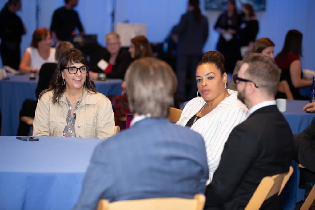 Four professionals sit around a round table in conversation at a networking event. Two women face the camera—one wearing glasses and a beige jacket, smiling as she speaks, and another in a white striped blazer listening attentively—while two men in suits sit with their backs partially to the camera. Other attendees and tables are visible in the softly lit background of the conference room.
