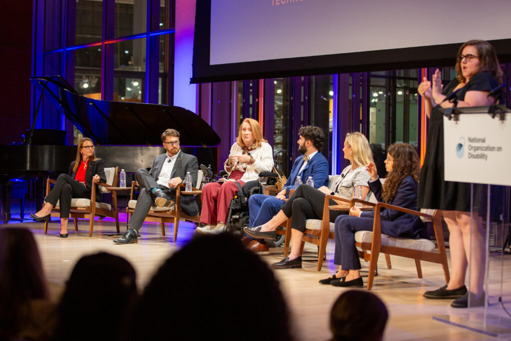 A photo of 6 people sitting on stage in conversation, with a person standing beside them translating the conversation into ASL.