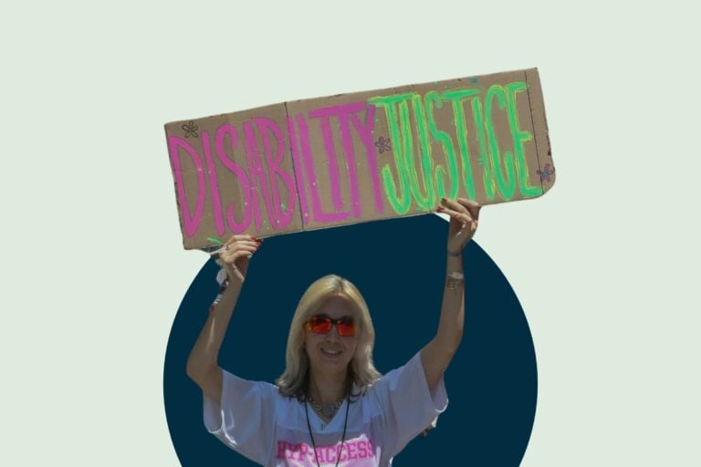 A person with blonde hair holds a cardboard sign reading "Disability Justice" in bright pink and green letters. They wear a white shirt and red sunglasses, smiling confidently.