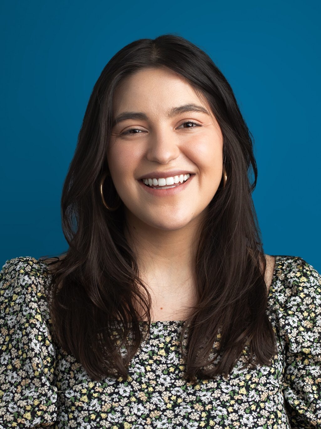 Smiling woman with long dark hair and hoop earrings, wearing a floral blouse against a blue background. The expression is warm and inviting.
