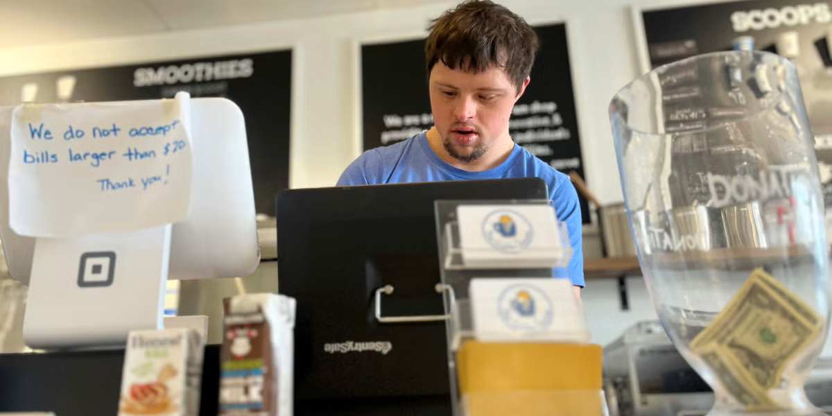 A person working at a counter in a café named "Scoops & Smoothies," standing next to a cash register and various beverages,