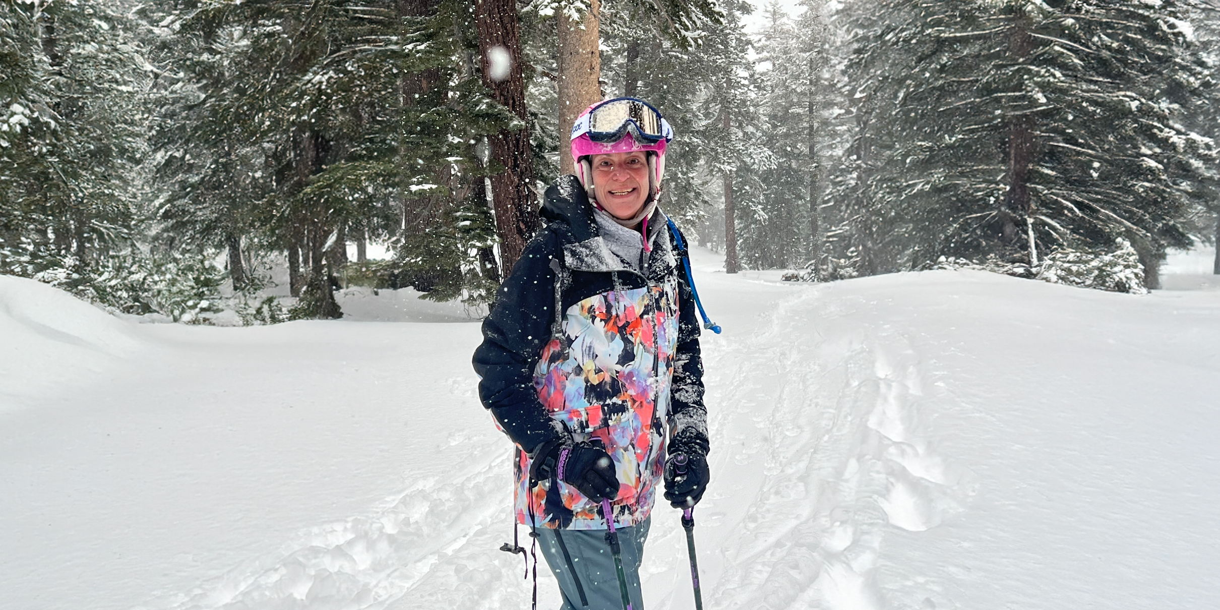 Beth Sirull wearing a helmet, winter jacket, and gloves, while holding onto ski poles. Behind Beth is a snow-covered forest.