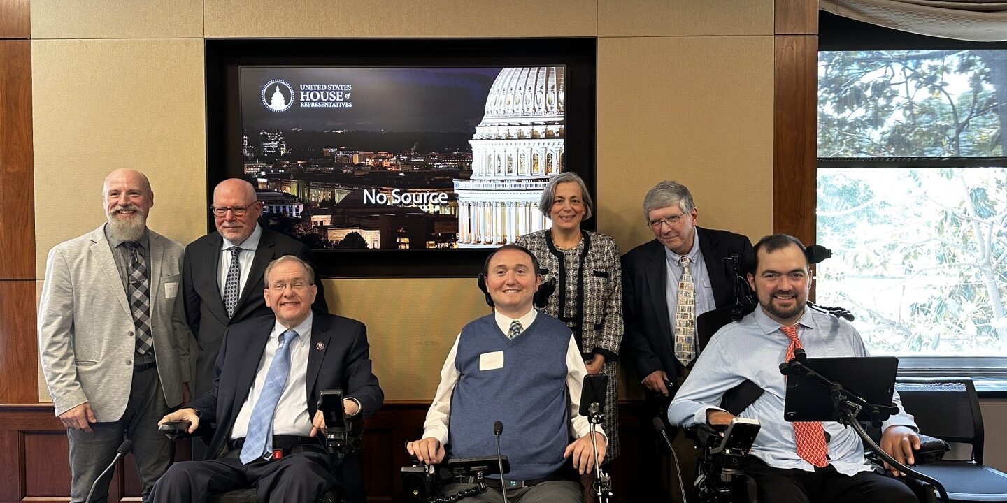 Image20250521105322 A group of seven people pose for a photo in a conference room at the U.S. House of Representatives. Three individuals are in power wheelchairs, while the others stand behind them. Everyone is smiling and dressed in business or professional attire. Behind the group is a large screen displaying an image of the U.S. Capitol and the House of Representatives logo. The setting suggests a formal meeting or advocacy event.