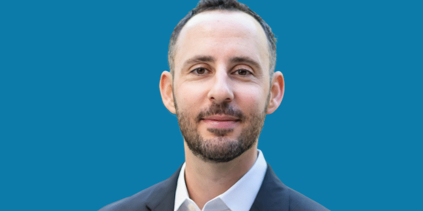 Headshot for Dan Robin, a white man with dark hair and a beard looking directly at the camera and smiling. He wears a white shirt and navy blazer. The background the NOD blue.