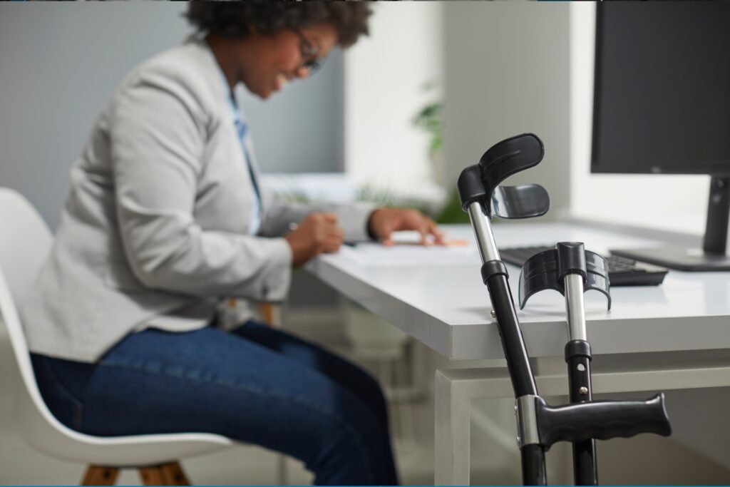 A person in a white blazer and blue pants sits at a white desk, writing or reviewing documents. Black forearm crutches are positioned against the desk nearby. The setting appears to be a modern office or workspace with a laptop visible in the background.