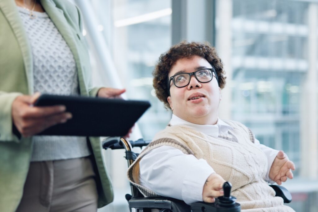 A person using a motorized wheelchair engages in conversation with a colleague holding a tablet in a modern office setting. They wear glasses, a white shirt, and a beige sweater vest, and appear confident and engaged.