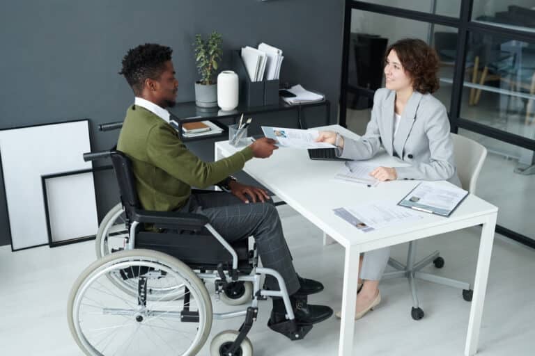 A woman sits behind a desk across from a man handing her his resume. He is seated in a wheelchair.