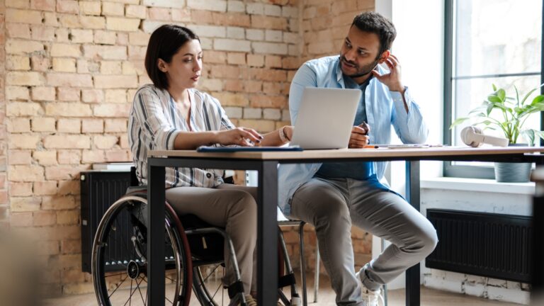 Two colleagues working together at a table, one using a wheelchair. They are focused and engaged, with a laptop and documents in a modern office.