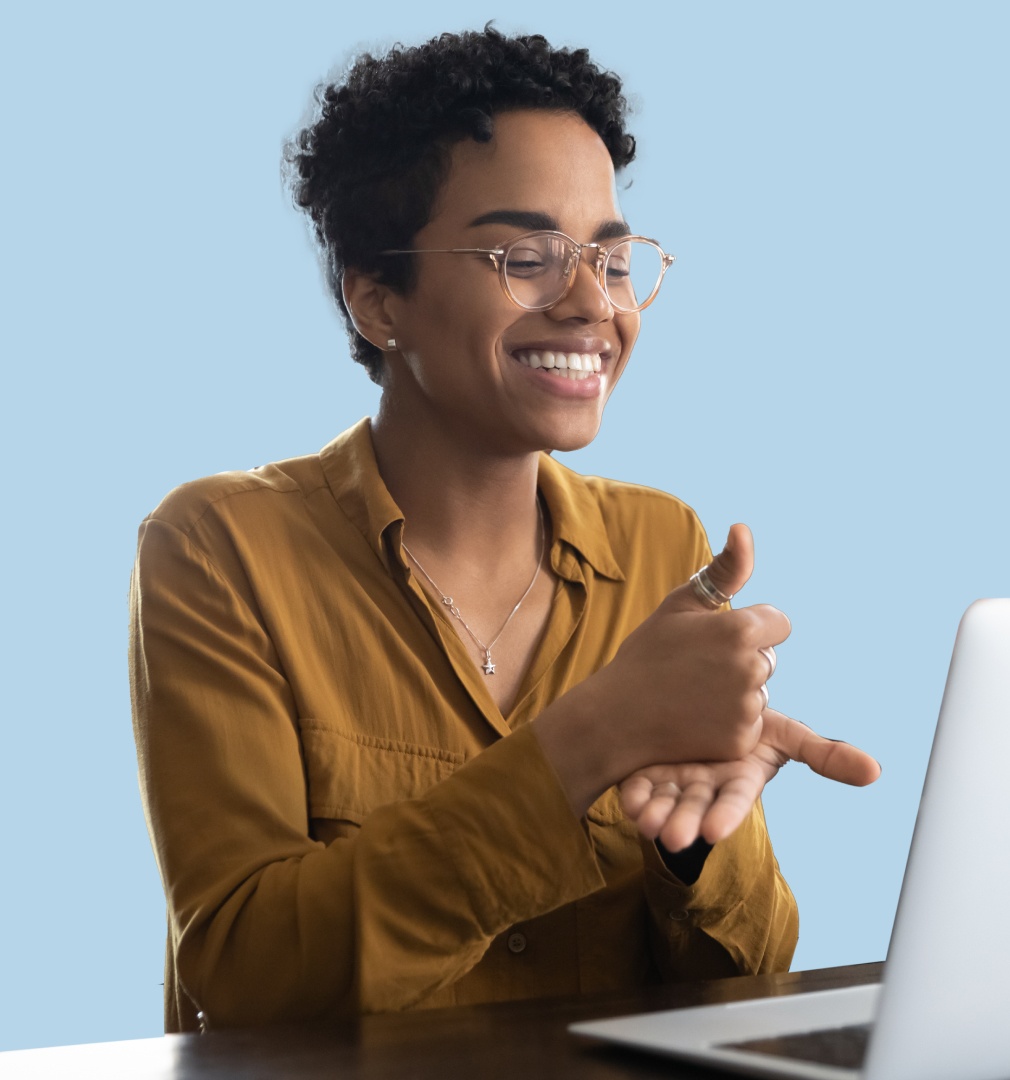 Professional woman with short curly hair and clear-framed glasses smiling and giving thumbs up gesture while working on laptop against light blue background"Detailed version: "Happy professional woman with short dark curly hair wearing clear-framed glasses and mustard yellow button-up shirt, smiling broadly while giving enthusiastic thumbs up with her right hand as she works on a laptop computer against a soft blue background