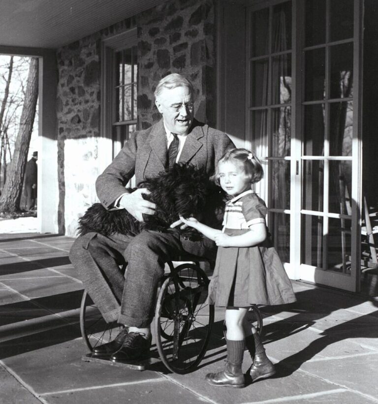 A black-and-white photo of President Franklin D. Roosevelt sitting in a wheelchair on a porch, holding a small black dog and smiling at a young girl standing beside him. A person is faintly visible in the background through the doorway.