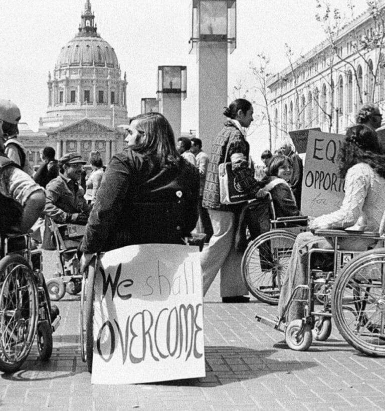 A black-and-white image of disability rights activists in wheelchairs gathered in front of San Francisco City Hall during the 504 Sit-in, holding protest signs including one that reads “We Shall Overcome” and another calling for equal opportunity.