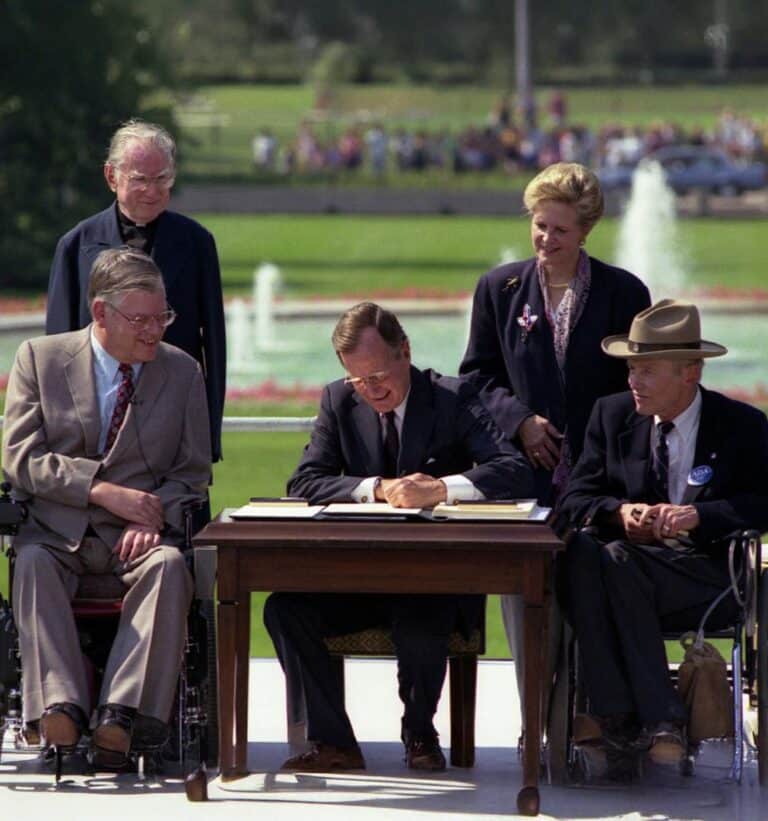 President George H. W. Bush signs the Americans with Disabilities Act into law on the White House lawn, seated at a table between two disability rights advocates in wheelchairs, with supporters standing behind them and a fountain in the background.
