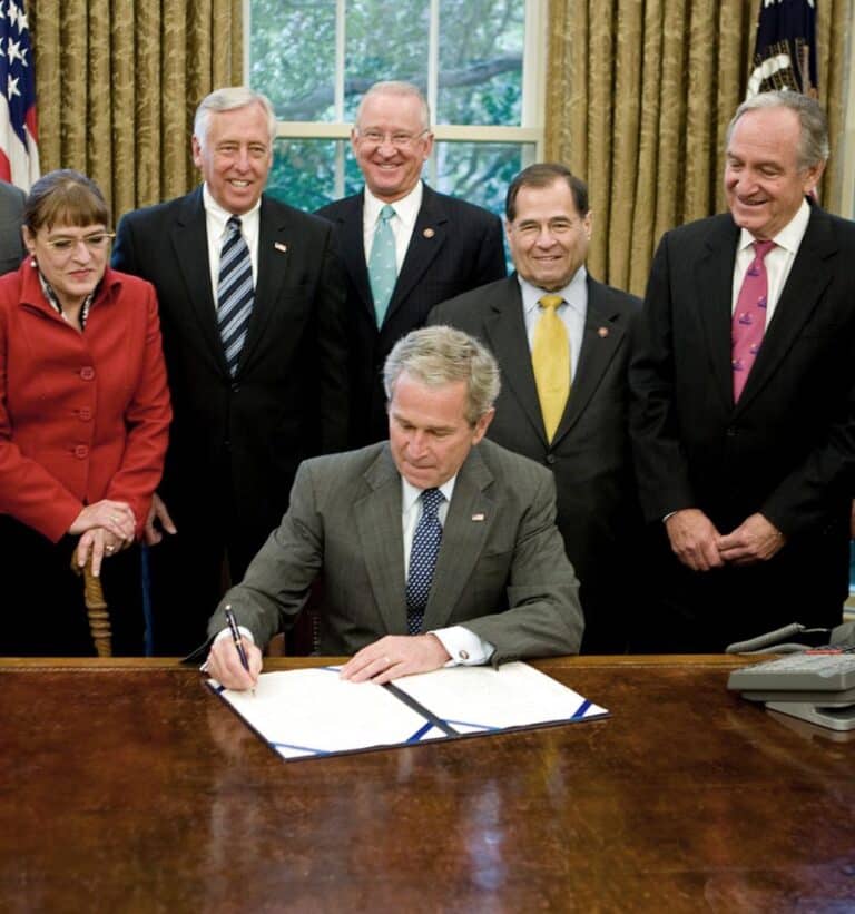 President George W. Bush sits at a desk in the Oval Office, signing a document surrounded by six smiling members of Congress, including both men and women, during a formal bill-signing ceremony.