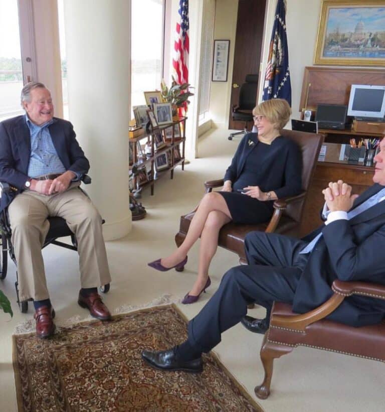 Former President George H. W. Bush sits in a wheelchair, smiling during a casual meeting in an office with former NOD President Carol Glazer and NOD Chairman Emeritus, Governor Tom Ridge, surrounded by personal photos, an American flag, and presidential memorabilia.