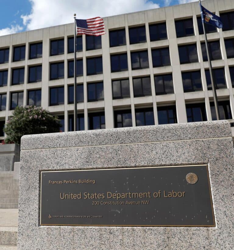 A daytime photo of the Frances Perkins Building, headquarters of the U.S. Department of Labor in Washington, D.C., with the American flag and Department of Labor sign prominently displayed in the foreground.