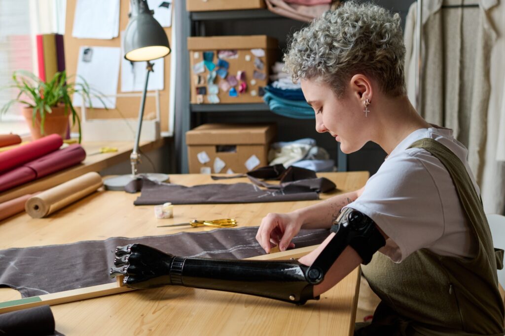 A person with short curly gray hair and earrings works at a wooden table in what appears to be a leather crafting workshop. They are focused on crafting or repairing a black leather item, with various tools and colorful fabric rolls visible in the background. The workspace includes shelving with materials, a desk lamp, and a small potted plant, creating an organized artisan's studio environment.