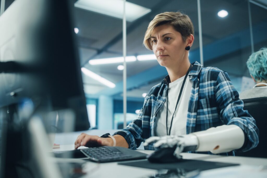A focused woman with short hair and a prosthetic arm works at a desktop computer in a modern office setting. She wears a blue plaid shirt over a white t-shirt and a lanyard, indicating a professional or tech work environment.