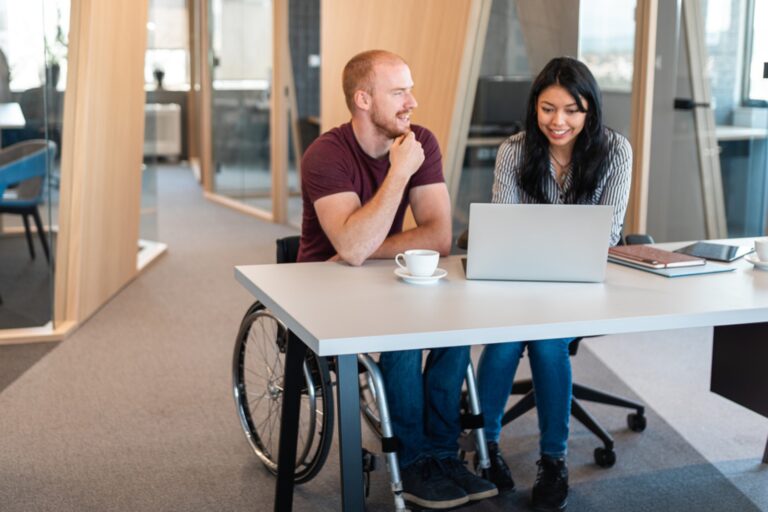 A man in a wheelchair wearing a burgundy polo shirt sits at a modern white desk with a woman in a black and white striped blouse, both smiling as they work together on a laptop. The man has his hand thoughtfully positioned near his chin while looking at the screen. The contemporary office space features large windows, wooden accents, and blue chairs in the background. A white coffee cup, notebooks, and a smartphone are visible on the desk, creating a collaborative and accessible workplace environment.