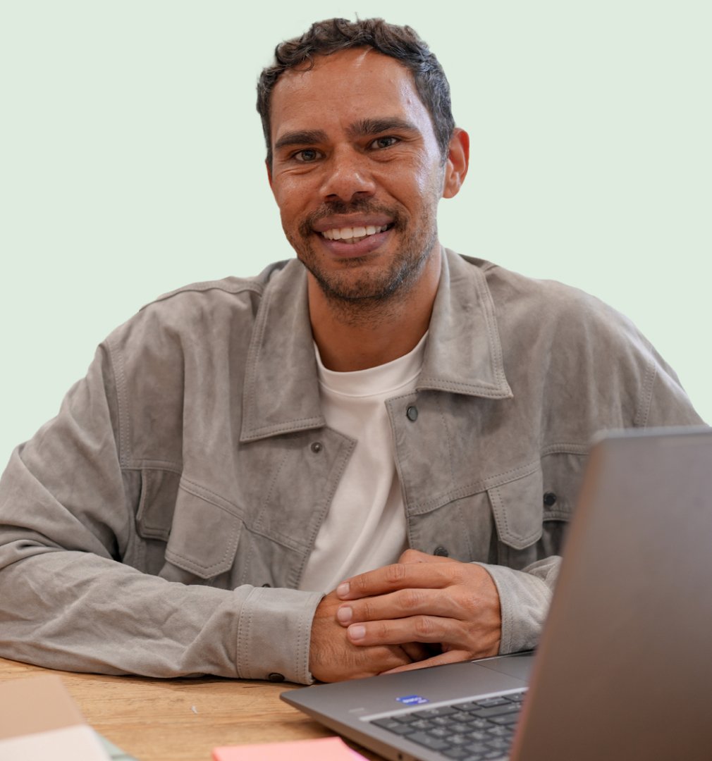 Smiling man in a gray jacket sits at a table with a laptop. The background is plain, conveying a friendly and casual atmosphere.