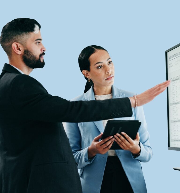 A standing man and woman looking at chart data on a computer monitor. The woman is holding a tablet and both are engaged in coversation