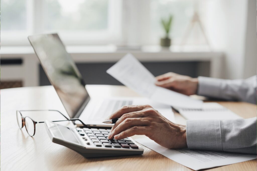A person in a light-colored dress shirt works at a wooden desk, with one hand typing on a calculator and the other holding a white document or paper. A laptop computer and eyeglasses are visible on the desk, along with scattered papers. The bright, modern office setting features large windows and a small potted plant in the blurred background, suggesting a professional accounting or financial work environment.