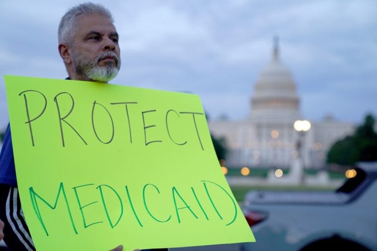 A man with gray hair and beard stands in front of the blurred U.S. Capitol building, holding a large bright yellow-green sign with the handwritten words “PROTECT MEDICAID” in bold black and green letters. The scene appears to be during dusk.