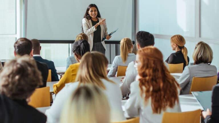 A woman in professional attire passionately presents to a seated audience in a bright conference room. The mood is engaging and focused.