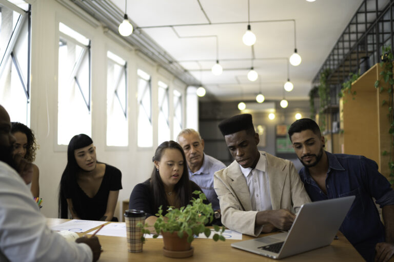 A diverse group of colleagues sit around a table in a modern office, collaborating and looking at a laptop. A woman with Down syndrome is actively engaged in the discussion as a man points at the screen.